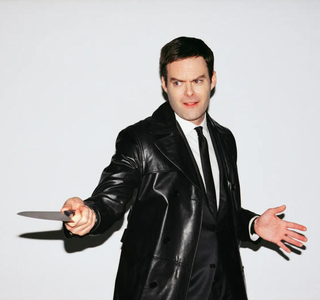 bill hader in a suit and leather overcoat, holding a knife, posed as if to jab it at someone