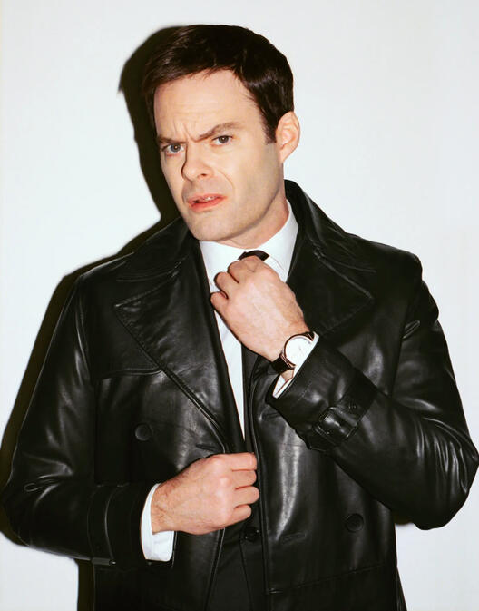 bill hader in a suit and leather overcoat, adjusting his tie.